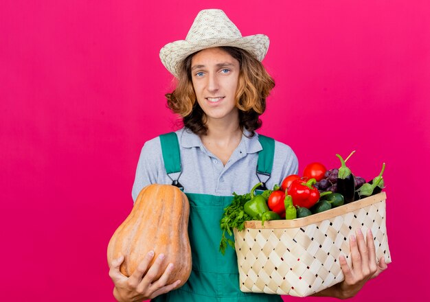 Jeune homme de jardinier portant une combinaison et un chapeau tenant une caisse pleine de légumes frais