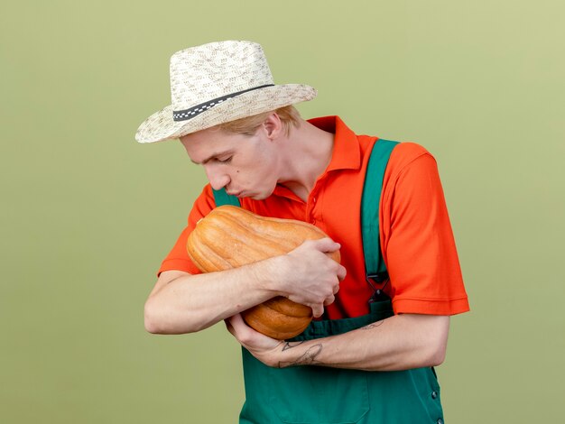 Jeune homme de jardinier portant combinaison et chapeau tenant la citrouille en le regardant embrasser debout sur fond clair