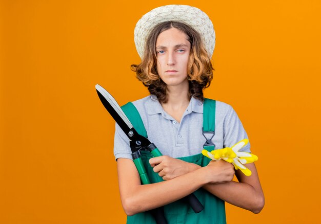 Jeune homme de jardinier portant combinaison et chapeau tenant un coupe-haie et des gants en caoutchouc