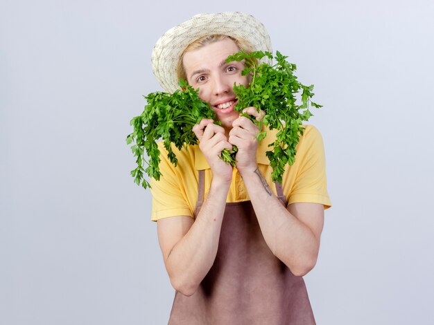 Jeune homme jardinier portant une combinaison et un chapeau tenant des herbes fraîches avec un sourire sur un visage heureux