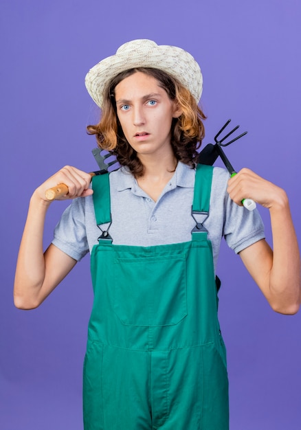 Jeune homme de jardinier portant combinaison et chapeau tenant un mini râteau et mattock