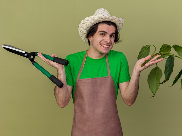 Jeune homme jardinier en tablier et chapeau tenant des tondeuses à plantes et haies regardant la caméra en souriant avec un visage heureux debout sur fond clair