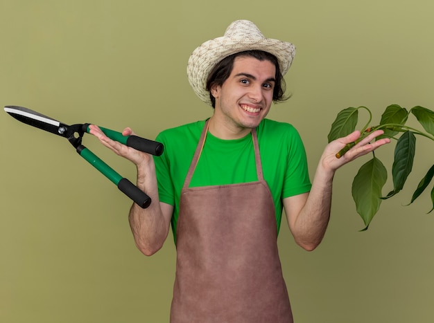 Jeune homme jardinier en tablier et chapeau tenant des tondeuses à plantes et haies regardant la caméra en souriant avec un visage heureux debout sur fond clair