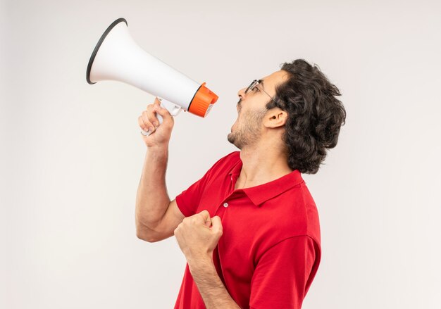 Jeune homme joyeux en chemise rouge avec des lunettes optiques crie par haut-parleur et regarde à côté isolé sur mur blanc