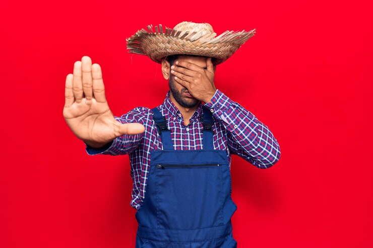 Photo gratuit jeune homme latin portant un chapeau de fermier et un tablier couvrant les yeux avec les mains et faisant un geste d'arrêt avec une expression de tristesse et de peur. concept embarrassé et négatif.