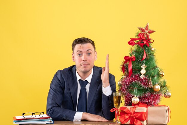 Jeune homme levant la main assis à la table près de l'arbre de Noël et présente sur jaune