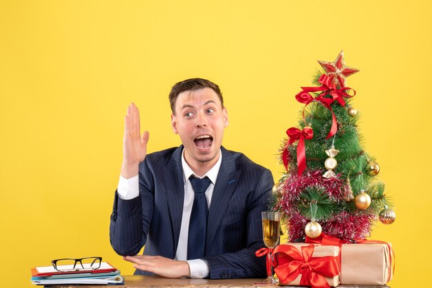 Jeune homme levant la main assis à la table près de l'arbre de Noël et présente sur jaune