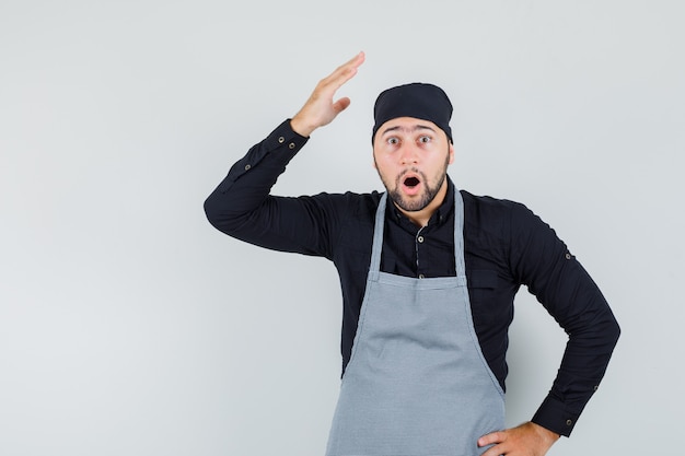 Jeune homme levant la main en chemise, tablier et à la vue choquée, de face.