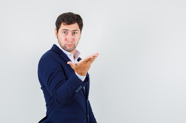 Jeune homme levant la main en geste indigné en costume bleu, vue de face.