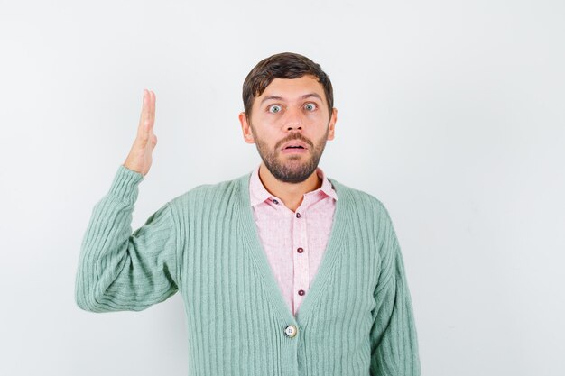 Jeune homme levant la main de manière perplexe en chemise, cardigan et l'air choqué. vue de face.