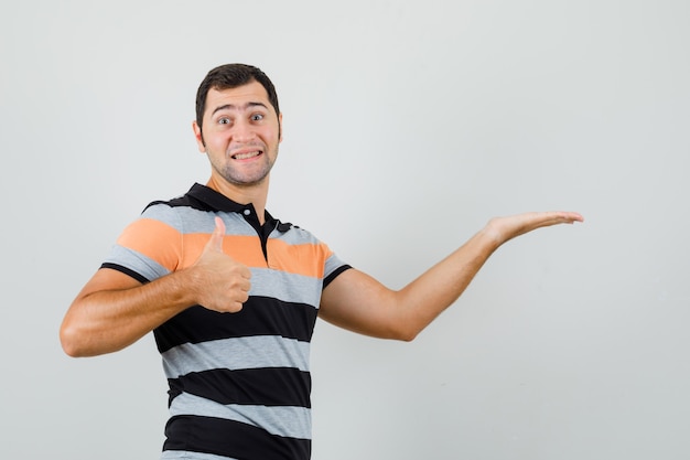 Jeune homme levant la main pour montrer quelque chose tout en montrant le pouce vers le haut en t-shirt et à la joyeuse. espace pour le texte