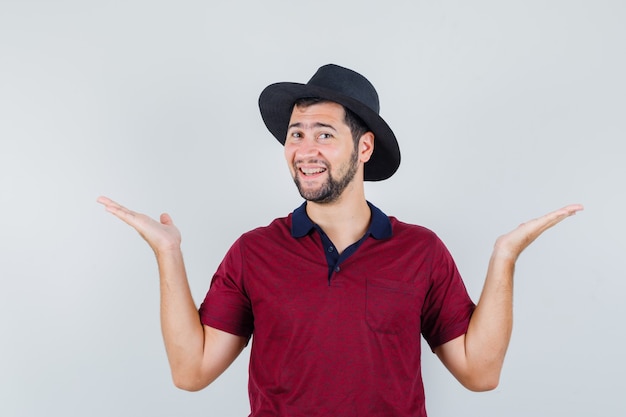 Jeune homme levant les mains comme tenant ou montrant quelque chose en t-shirt, chapeau et à la joyeuse. vue de face.