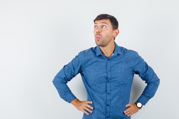 Jeune homme levant les mains sur la taille en chemise bleue et l'air pensif. vue de face.