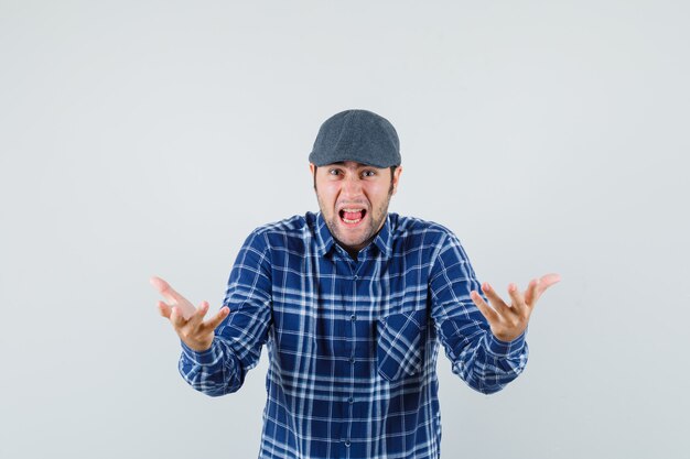 Jeune homme levant les mains tout en criant en chemise, casquette et à la colère, vue de face.