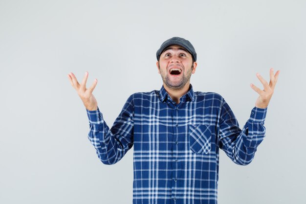 Jeune homme levant les mains tout en regardant en chemise, casquette et à la reconnaissance. vue de face.