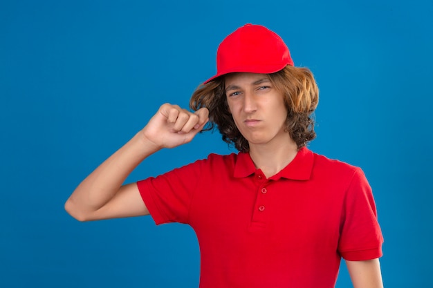 Jeune homme de livraison bouleversé en uniforme rouge touchant ses cheveux regardant la caméra montrant l'aversion sur fond bleu isolé