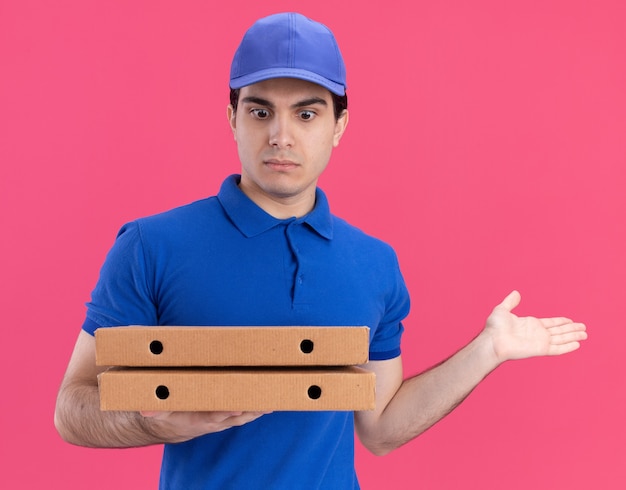 Jeune homme de livraison caucasien impressionné en uniforme bleu et casquette tenant et regardant des paquets de pizza montrant la main vide