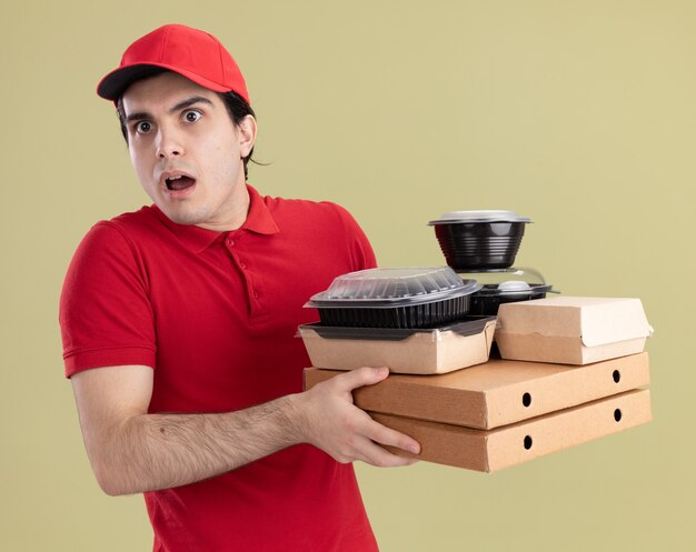 Jeune homme de livraison caucasien impressionné en uniforme rouge et casquette tenant des emballages de pizza avec des contenants de nourriture et des emballages de nourriture en papier sur eux en regardant de côté