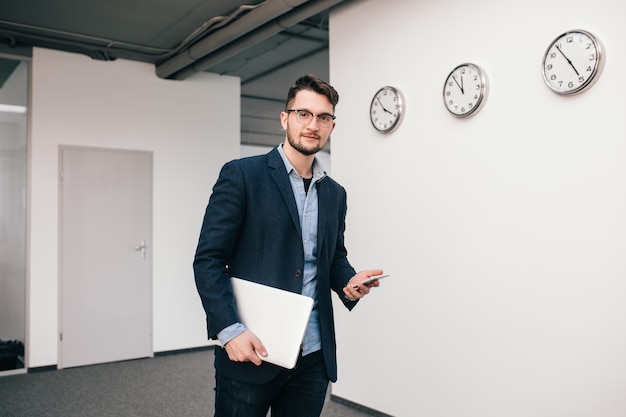 Jeune homme à lunettes marche au bureau. Il porte une chemise bleue, une veste sombre, un jean et une barbe. Il tient le téléphone et l'ordinateur portable dans les mains. Regardant la caméra.