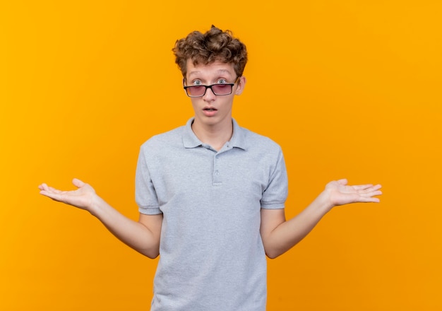 Jeune homme à lunettes noires portant un polo gris à la confusion et surpris en levant les bras s'étendant sur les côtés n'ayant pas de réponse debout sur un mur orange