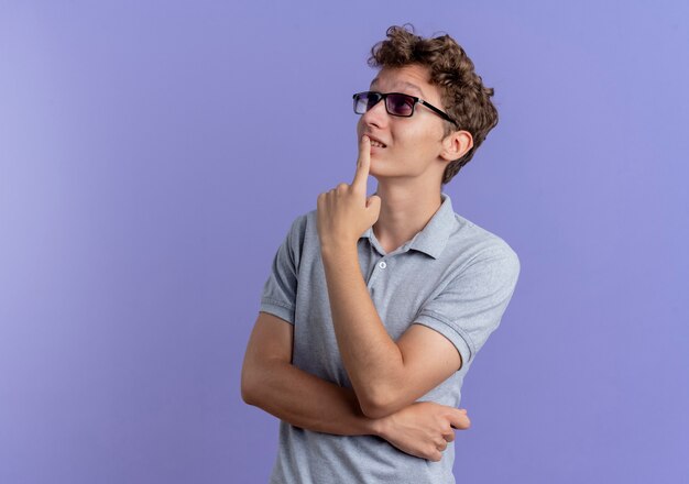 Jeune homme à lunettes noires portant un polo gris à côté avec une expression pensive pensée positive souriant sur bleu