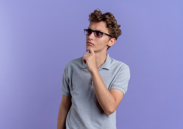 Jeune homme à lunettes noires portant un polo gris à côté perplexe debout sur le mur bleu