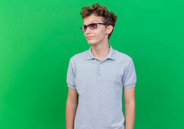 Jeune homme à lunettes noires portant un polo gris à côté avec le sourire sur le visage debout sur un mur vert