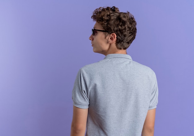 Jeune homme à lunettes noires portant un polo gris debout avec son dos sur un mur bleu