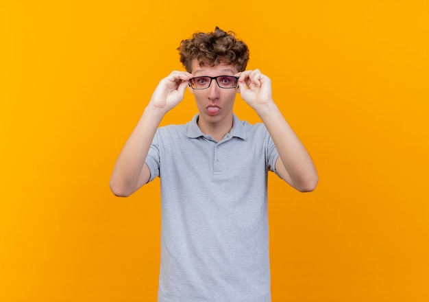 Jeune homme à lunettes noires portant un polo gris faisant grimace qui sort la langue debout sur un mur orange