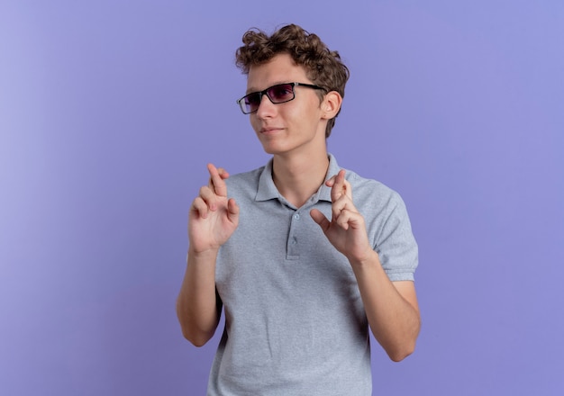 Jeune homme à lunettes noires portant un polo gris faisant souhait souhaitable croisant les doigts debout sur le mur bleu