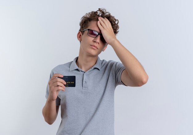 Jeune homme à lunettes noires portant un polo gris montrant une carte de crédit à la confusion et anxieux debout sur un mur blanc