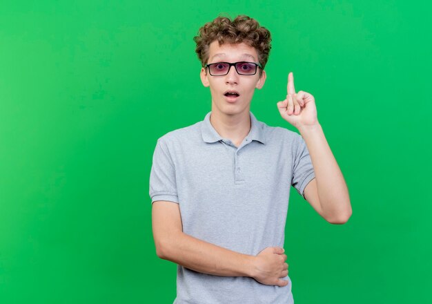 Jeune homme à lunettes noires portant un polo gris montrant l'index d'être surpris et heureux d'avoir une nouvelle idée debout sur un mur vert