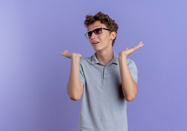 Jeune homme à lunettes noires portant un polo gris regardant de côté confus écartant les bras sur les côtés debout sur le mur bleu