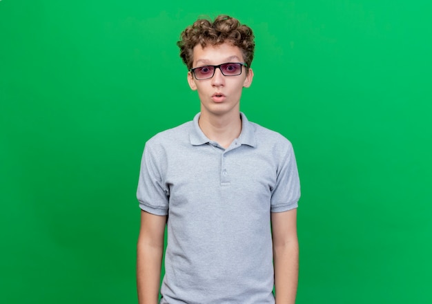 Jeune homme à lunettes noires portant un polo gris surpris et étonné debout sur le mur vert