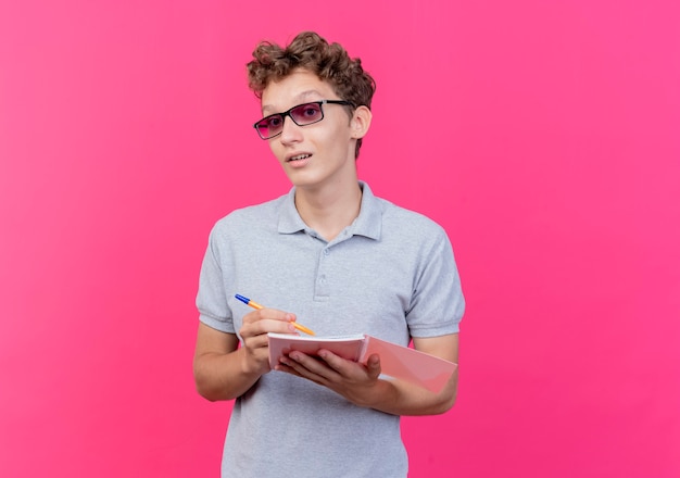 Jeune homme à lunettes noires portant un polo gris tenant un cahier avec un stylo heureux et positif souriant sur rose