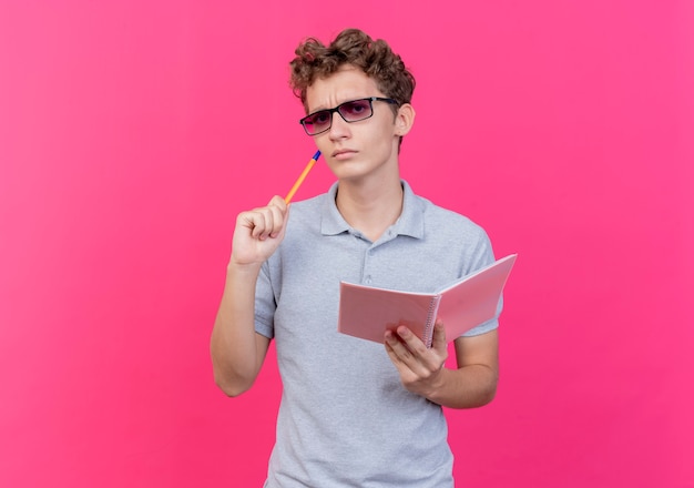 Jeune homme à lunettes noires portant un polo gris tenant un cahier avec un stylo à la réflexion avec une expression pensive sur rose