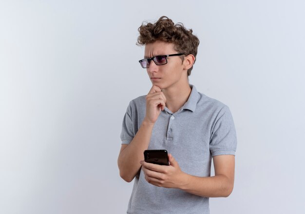 Jeune homme à lunettes noires portant un polo gris tenant le smartphone à côté avec la main sur le menton avec une expression pensive debout sur un mur blanc