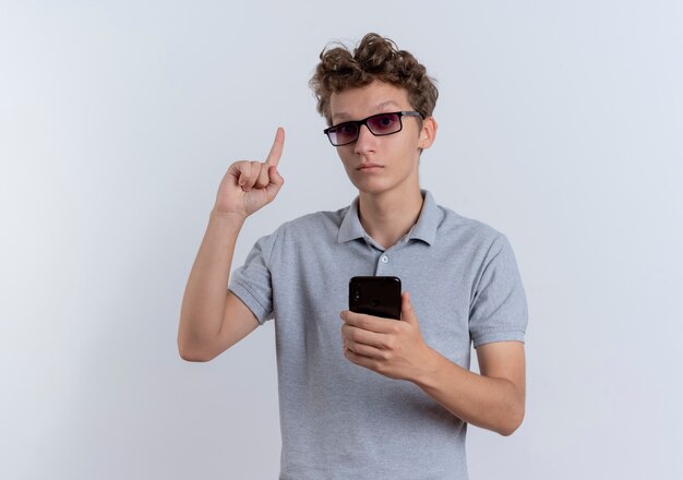 Jeune homme à lunettes noires portant un polo gris tenant un smartphone montrant l'index à la surprise d'avoir une nouvelle idée debout sur un mur blanc