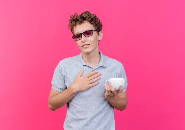 Jeune homme à lunettes noires portant un polo gris tenant une tasse de café avec la main sur la poitrine se sentant reconnaissant heureux et positif sur rose