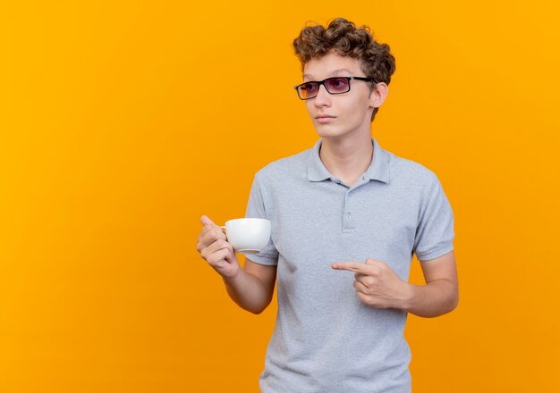 Jeune homme à lunettes noires portant un polo gris tenant une tasse de café pointant avec le doigt à la regarder de côté avec un visage sérieux debout sur un mur vert