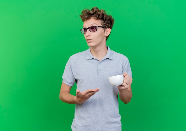 Jeune homme à lunettes noires portant un polo gris tenant une tasse de café présentant avec bras à côté avec une expression confiante sérieuse sur vert