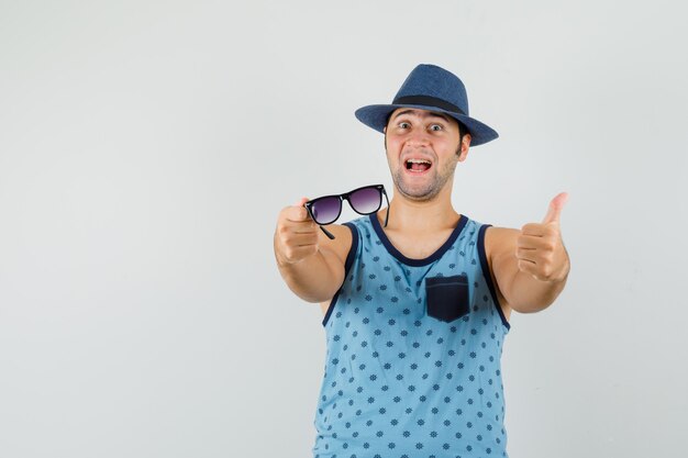 Jeune homme en maillot bleu, chapeau tenant des lunettes, montrant le pouce vers le haut et l'air heureux