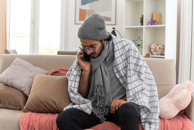 Photo gratuite jeune homme malade anxieux dans des lunettes optiques enveloppées dans un plaid avec une écharpe autour du cou portant un chapeau d'hiver parlant au téléphone assis sur un canapé dans le salon