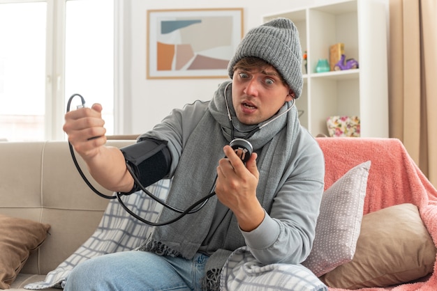 jeune homme malade anxieux avec une écharpe autour du cou portant un chapeau d'hiver enveloppé dans un plaid mesurant sa pression avec un sphygmomanomètre assis sur un canapé dans le salon