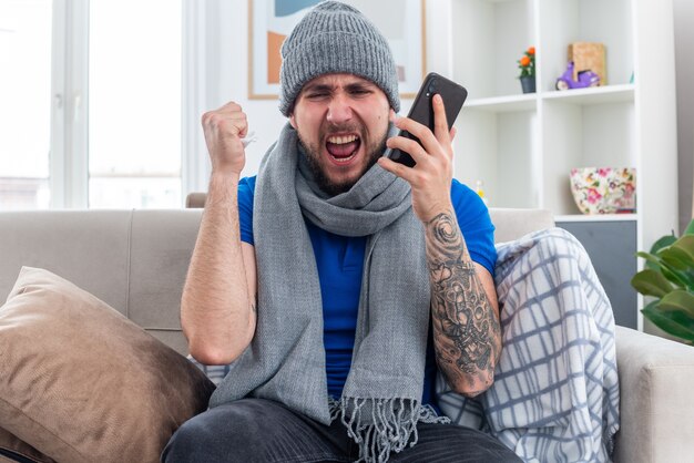 Jeune homme malade en colère portant une écharpe et un chapeau d'hiver assis sur un canapé dans le salon tenant une serviette et un téléphone portable regardant vers le bas en serrant le poing criant