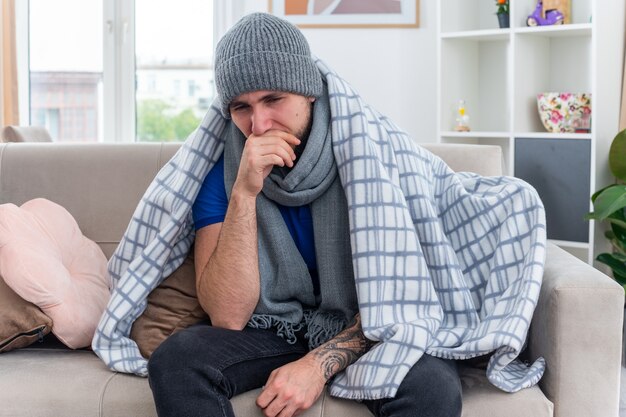 Jeune homme malade faible et douloureux portant une écharpe et un chapeau d'hiver enveloppé dans une couverture assis sur un canapé dans le salon regardant vers le bas en gardant la main sur la bouche