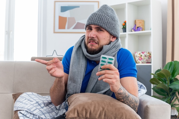 jeune homme malade irrité portant une écharpe et un chapeau d'hiver assis sur un canapé dans le salon avec un oreiller sur ses jambes tenant un paquet de capsules regardant l'avant pointant sur le côté