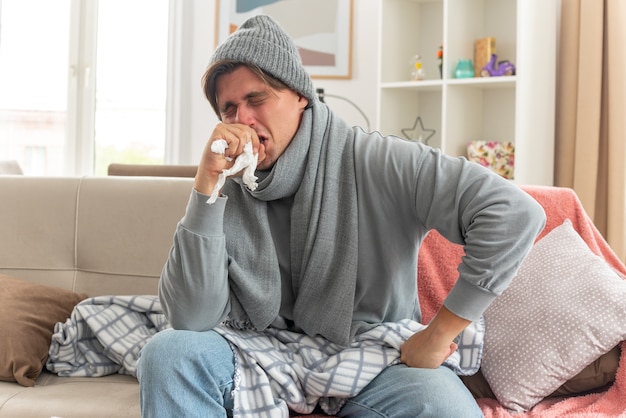 Jeune homme malade mécontent avec une écharpe autour du cou portant un chapeau d'hiver éternue tenant la main près du visage assis sur un canapé au salon