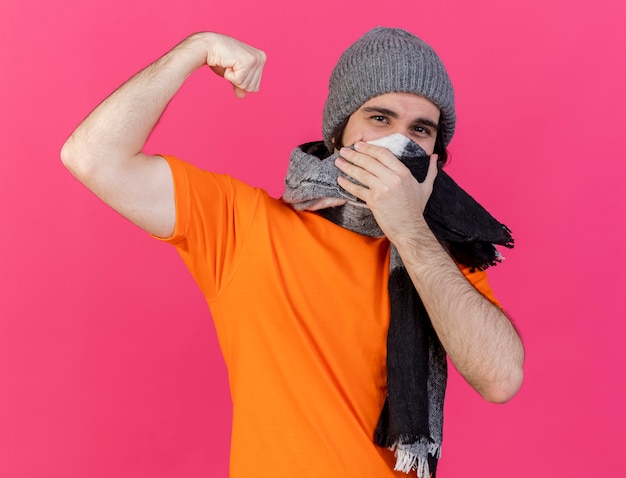 Photo gratuite jeune homme malade portant chapeau d'hiver avec écharpe montrant le visage couvert de geste fort avec foulard isolé sur rose