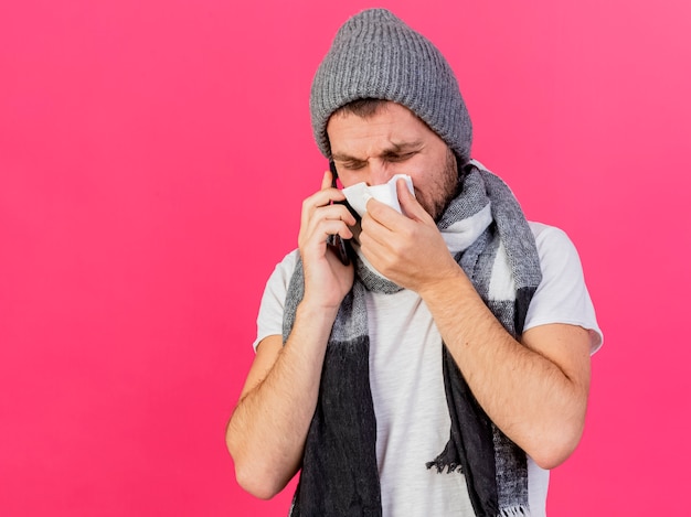 Jeune homme malade portant un chapeau d'hiver avec écharpe parle au téléphone et essuyant le nez avec une serviette isolé sur rose